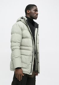 Pufferjakke i lys grå, med hætte, diagonale syninger, lynlås lukning og flere lommer, båret over en sort top.