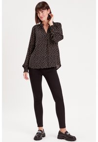 Black polka-dot blouse with long sleeves and buttoned cuffs, featuring a relaxed fit, paired with black skinny pants and chunky black shoes.