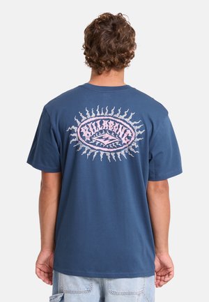 Uomo con capelli ricci che indossa una maglietta blu con una grafica rosa e bianca a raggi di sole e testo sul retro, abbinato a pantaloncini di jeans chiari.