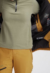 Osoba zakładająca musztardowożółtą kurtkę Gore-Tex na zielony polar O'Neill oraz pasujące żółte spodnie.