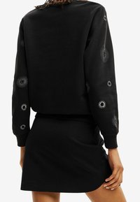 Czarny sweatshirt z haftowanymi spiralnymi wzorami na rękawach, w połączeniu z czarną spódnicą. Miękka tkanina; luźny krój i okrągły dekolt.