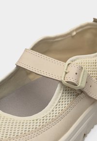 Beige Mary Jane-stil sko med mesh-overside, justerbar stropp og en plastspenne på hvit bakgrunn.