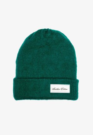 Zöld kötött sapka puha anyagból, hajtott passzéval. Az oldalán fekete felirattal ellátott fehér címke található.