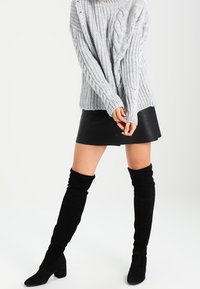 Grau gestrickter Pullover, schwarzer lederner Minirock und schwarze Stiefel aus Wildleder in Oberschenkellänge. Der Pullover hat eine gerippte Textur und eine oversized Passform.