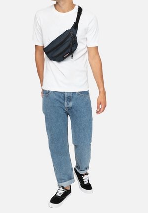 Marsupio in tessuto nero e blu scuro con cerniera, indossato a tracolla, dotato di una cintura regolabile, abbinato a una maglietta bianca e jeans chiari.