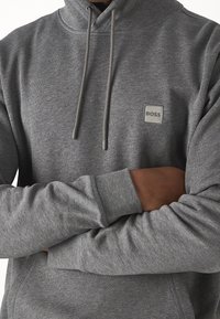 Gray hoodie with a textured fabric, drawstrings, and a small rectangular logo patch. Ribbed cuffs and a relaxed fit.