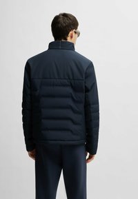 Marinblå puffjacka med hög krage, kviltad textur och blixtlås fram. Har långa ärmar med släta manschetter. Slätt, lättviktsmaterial.