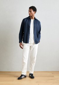 Granatowa koszula z guzikami, biały T-shirt, off-white spodnie i czarne sneakersy z białymi paskami. Naturalna drewniana podłoga i jasnoszare tło.