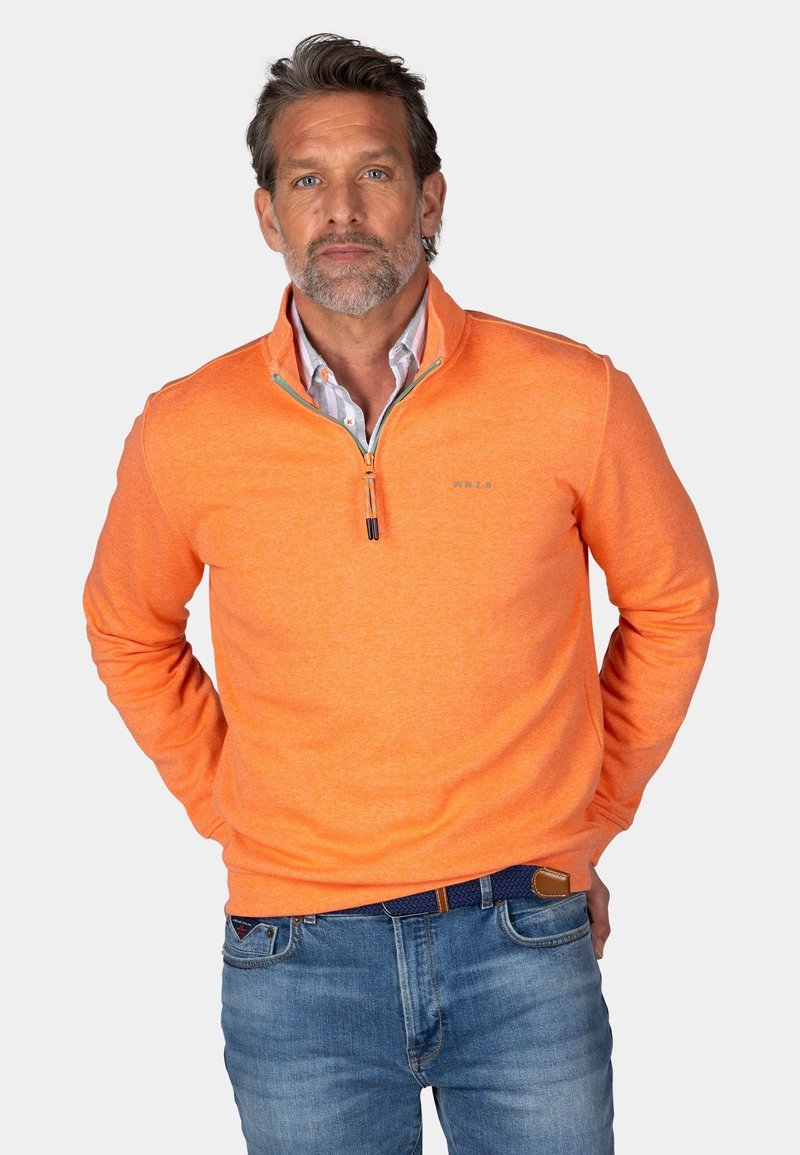 Pomarańczowy sweter z zamkiem błyskawicznym przy kołnierzu, wykonany z miękkiego materiału. Posiada ściągacze przy mankietach i u dołu, zestawiony z niebieskimi dżinsami.