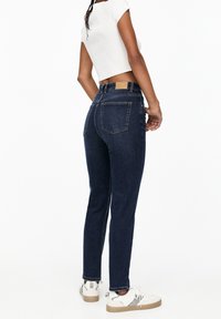 Jeans in denim blu scuro con una vestibilità alta, dotati di un yoke posteriore, due tasche posteriori e orlo grezzo, abbinati a sneakers bianche.