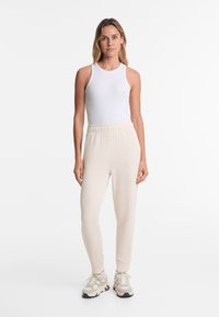 White sleeveless top paired with light beige jogger pants. Pants feature an elastic waistband and cuffed ankles. Sneakers are light-colored with accents.