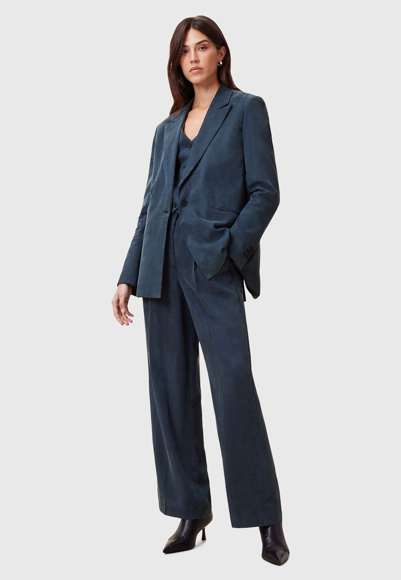 Completo blu scuro realizzato in tessuto morbido, composto da un blazer su misura e pantaloni a gamba larga, abbinato a décolleté nere con punta affilata.