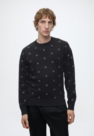 Young man with wavy light brown hair wearing a black sweater with small light grey "KL" patterns and black pants against a plain background.