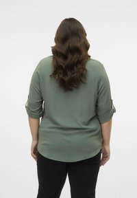 Blouse verte à texture avec des manches retroussées, vue de dos ; design semi-ajusté, ourlet arrondi, associée à un pantalon noir. Style simple et décontracté.