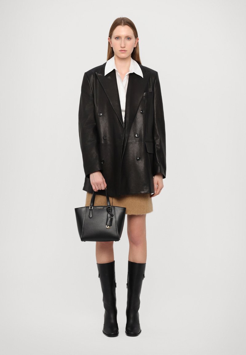 Blazer noir en cuir à double boutonnage, chemise blanche, jupe beige, bottes noires montantes et petit sac à main noir. Textures lisses et design ajusté.