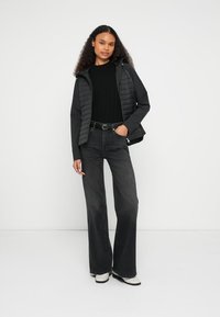 Black padded jacket layered over a black ribbed sweater, paired with high-waisted dark denim jeans. Model stands against a plain background.