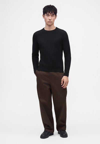 Jeune homme debout portant un pull noir à manches longues, un pantalon marron ample et des chaussures noires, sur un fond blanc uni.