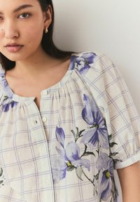 Blusa a fantasia floreale in crema con fiori viola e design a griglia. Presenta un colletto rotondo e una chiusura con bottoni, con dettagli di delicate pieghe.