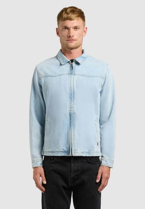 Ung mand iført en lyseblå lynlåstrøje i denim og sorte jeans, stående foran en ensfarvet lysegrå baggrund.