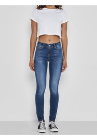 Jeans ajustados azul oscuro con una cintura alta, que cuentan con bolsillos delanteros y cierre de botón. Combinados con una camiseta blanca corta y zapatillas negras.