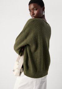 Stickad olivgrön tröja med lös passform och vida ärmar, kombinerad med en ljusbeige crossbody-väska. Strukturerad tyg och subtil detalj på baksidan.