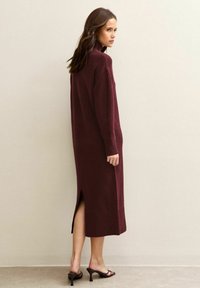 Donna in piedi di lato, che indossa un lungo vestito di maglia bordeaux con uno spacco posteriore e sandali neri con tacco, davanti a un muro beige semplice.