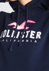 Mörkblå huvtröja med rosa mås-logotyp och vit text "Hollister California" på bröstet, snoddar hängande ned.