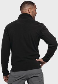Schwarzer Fleece-Pullover mit hohem Kragen, langen Ärmeln und glatter Textur, kombiniert mit grauen Hosen. Die Ansicht von hinten zeigt das taillierte Design.