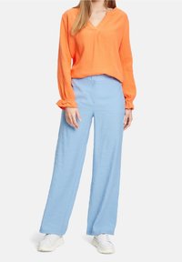 Blusa arancione a maniche lunghe con scollatura a V e tessuto strutturato, abbinata a pantaloni larghi blu chiaro e sneakers bianche.