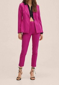 Kobieta w jaskraworóżowym, dopasowanym garniturze z krótkimi spodniami, czarną krótko przyciętą koszulą i czarnych sandałkach na cienkich paskach na jednolitym beżowym tle.