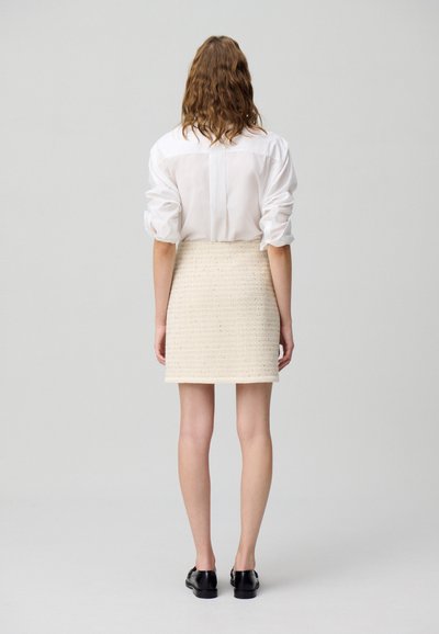 Chemise blanche à manches retroussées, associée à une jupe texturée crème. Des chaussures noires viennent compléter la tenue, avec un design minimaliste.