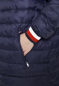 Námořnicky modrá prošívaná bunda s boční kapsou na zip. Manžety mají lem s červenými, bílými a námořnicky modrými pruhy. Hladká, prošitá textura.