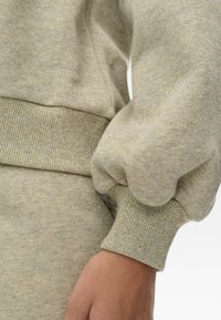 Sudadera gris claro con tejido texturizado y destellos dorados, puños acanalados y un corte relajado. Enfócate en el detalle de la manga y el material.