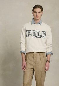Sweatshirt alb cu logo "POLO" în culoare marină, purtat peste o cămașă cu dungi albastre și albe, asortat cu pantaloni bej în carouri.