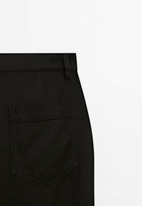 Gros plan sur un tissu noir montrant la poche arrière et la passants de ceinture d’un pantalon sur fond blanc.