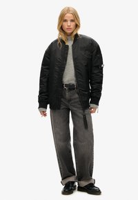 Sort polstret bomberjakke med ribbede mansjetter, grå sweater, sort bælte og brede grå jeans. Kraftige sorte sko fuldender outfittet.