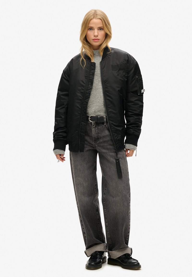 Sort polstret bomberjakke med ribbede mansjetter, grå sweater, sort bælte og brede grå jeans. Kraftige sorte sko fuldender outfittet.