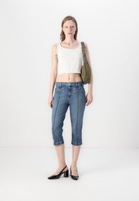 Top bianco tagliato, jeans capri in denim blu e tacchi in vernice nera. La modella porta una borsa a spalla verde. Stile semplice e moderno.