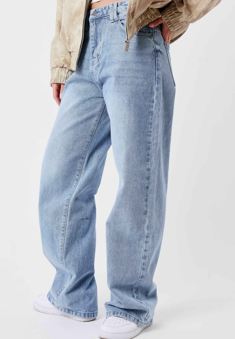 Personne portant un jean bleu clair à jambes larges, une veste beige et des baskets blanches, debout avec une main posée sur la hanche.