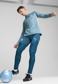 Completo da allenamento Puma in azzurro chiaro e teal. Maglia a maniche lunghe con logo; pantaloni aderenti con un motivo testurizzato. Inclusi scarpe nere e pallone.