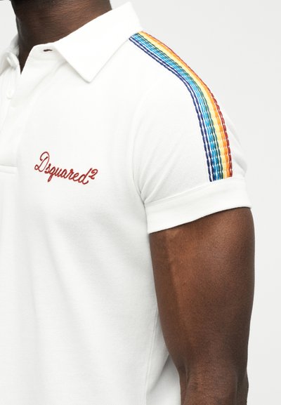 Gros plan sur une personne portant un polo blanc avec le logo « Dsquared² » brodé en rouge et une couture à rayures arc-en-ciel sur l'épaule.
