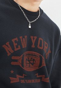 Sudadera negra con la palabra "NUEVA YORK" y un gráfico de fútbol en rojo desgastado. Se lleva un collar de cadena plateada con un colgante en forma de candado.