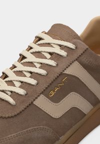 Καφέ σουέτ αθλητικό παπούτσι με μπεζ δερμάτινες λεπτομέρειες, λευκά κορδόνια και χρυσό λογότυπο "GANT" στο πλάι, με λαστιχένια σόλα.