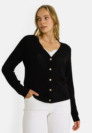 Cárdigan negro con cuello en V, con un patrón texturizado y botones dorados. Llevado sobre una blusa blanca con cuello de encaje y combinado con pantalones blancos.