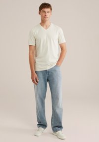 Krémové tričko s výstřihem do V v kombinaci s světle modrými džíny rovného střihu. Modelka stojí vklidu v bílých teniskách na neutrálním pozadí.