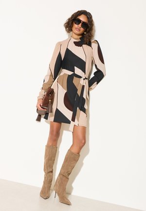 Sieviete, kas valkā saulesbrilles, rakstainu bēša, melnas un brūnas krāsas kleitu ar jostu, bēšas zamša augstās zābaku līdz ceļiem un rokās tur brūnu ādas somiņu ar bārkstīm.