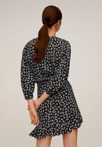 Schwarzes Kleid mit weißem Blumenmuster, lange Ärmel mit elastischen Bündchen und rüschenbesäumtem Saum, von hinten betrachtet.