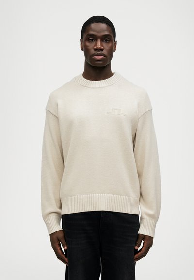 Beige knit sweater with a round neck, ribbed cuffs and hem, featuring a small, textured logo on the chest. Worn with dark jeans.