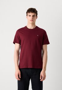 Bordeaux rundhalset t-shirt lavet af bomuld, med korte ærmer og et lille logo på brystet, parret med mørke denimbukser.