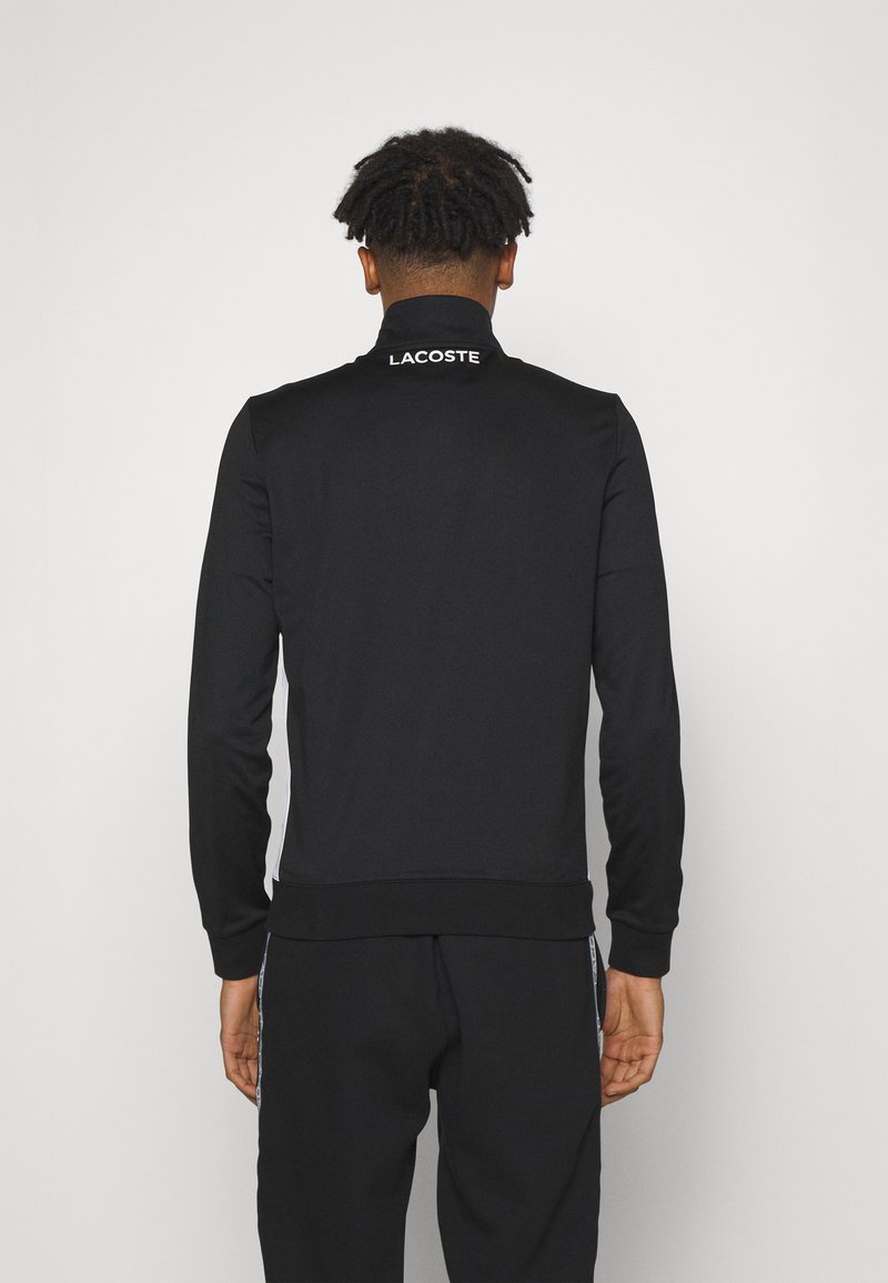 Zwarte sportjack met rits en hoge kraag. Bevat een wit "LACOSTE" logo bij de hals, gemaakt van een gladde, rekbare stof.